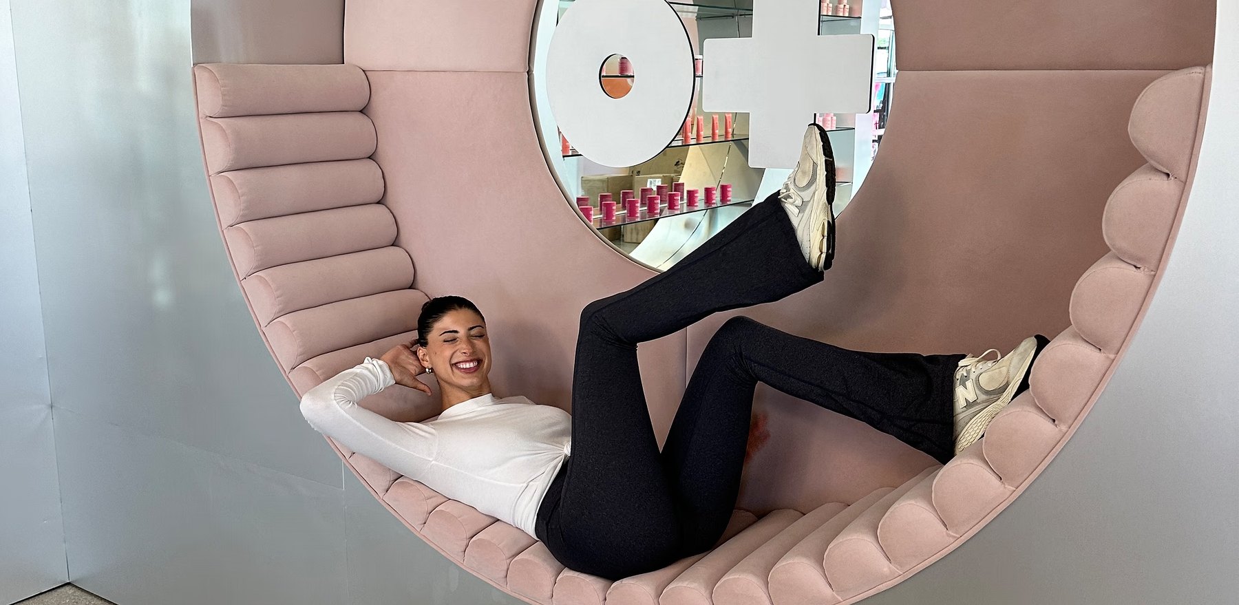 Brianna Bitton lounges on curved pink seating in a store.